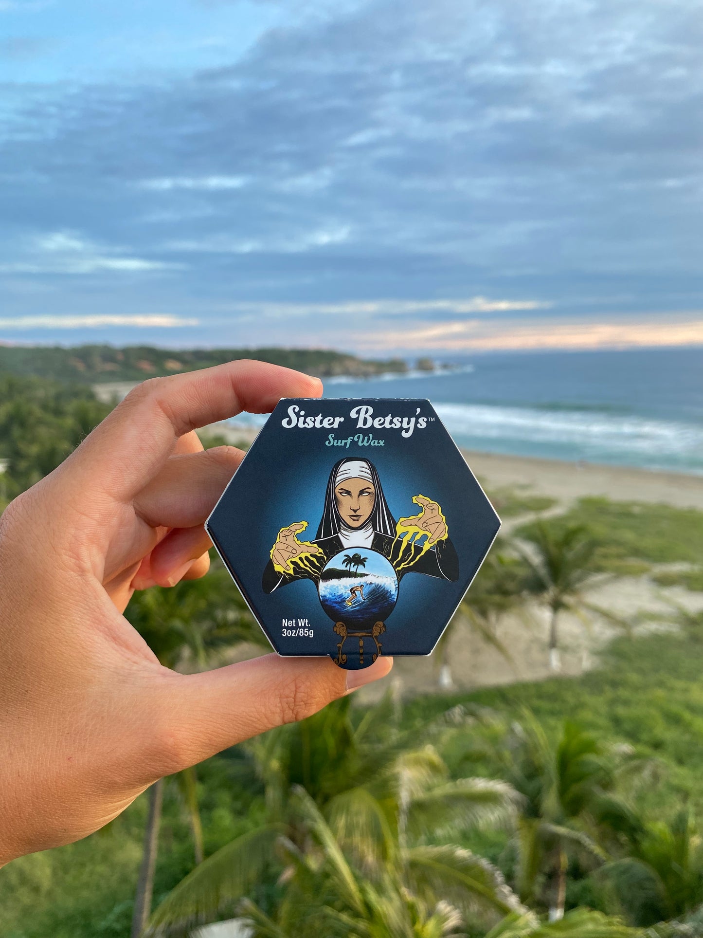 Hand holding a block of Sister Betsy's surfwax with a scenic beach in the background.